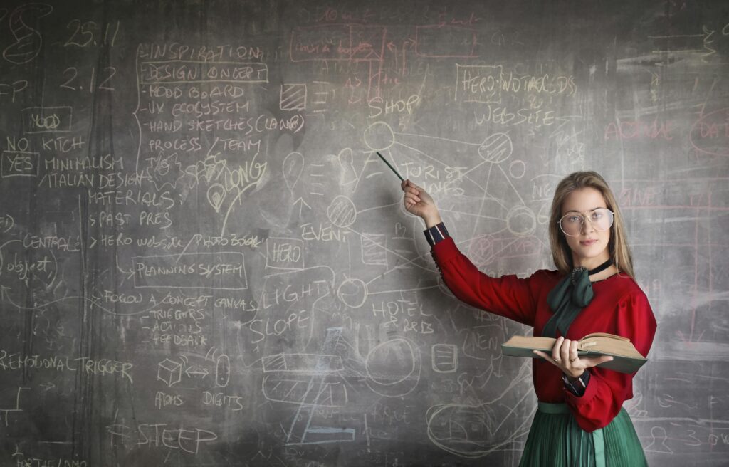 Dumbest Financial Advice, Serious female teacher wearing old fashioned dress and eyeglasses standing with book while pointing at chalkboard with schemes and looking at camera